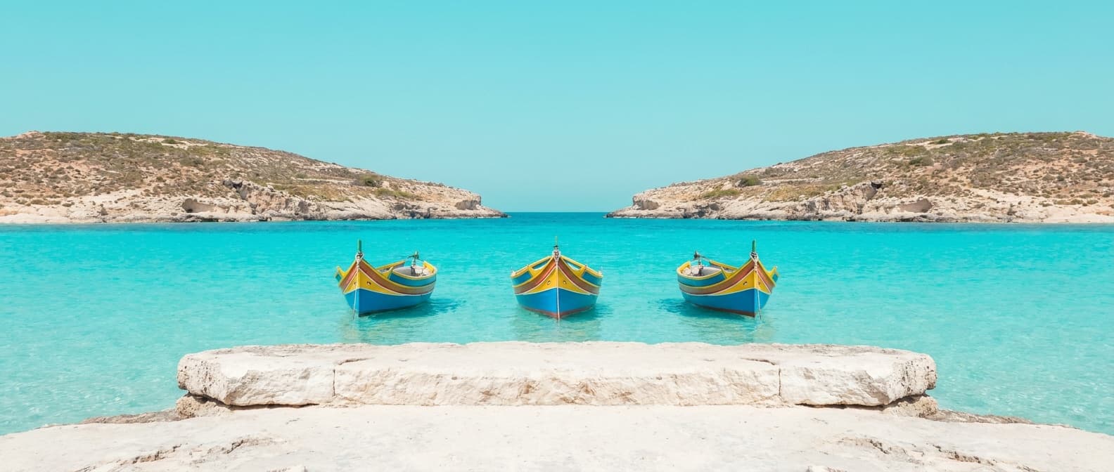 The Blue Lagoon at Comino — three painted luzzu boats at anchor in a row over bright turquoise shallows