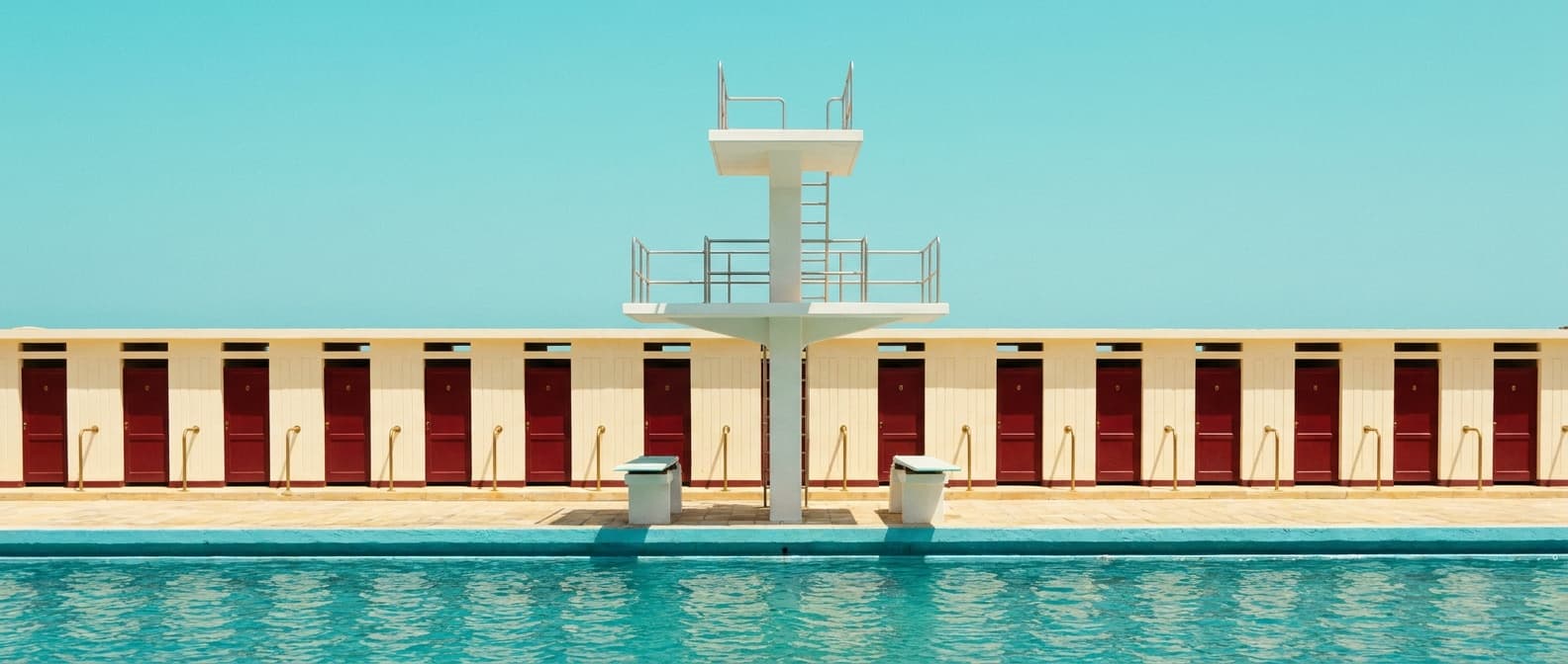 A symmetrical white three-tier diving platform over a calm turquoise sea, with cream changing cabins along the promenade